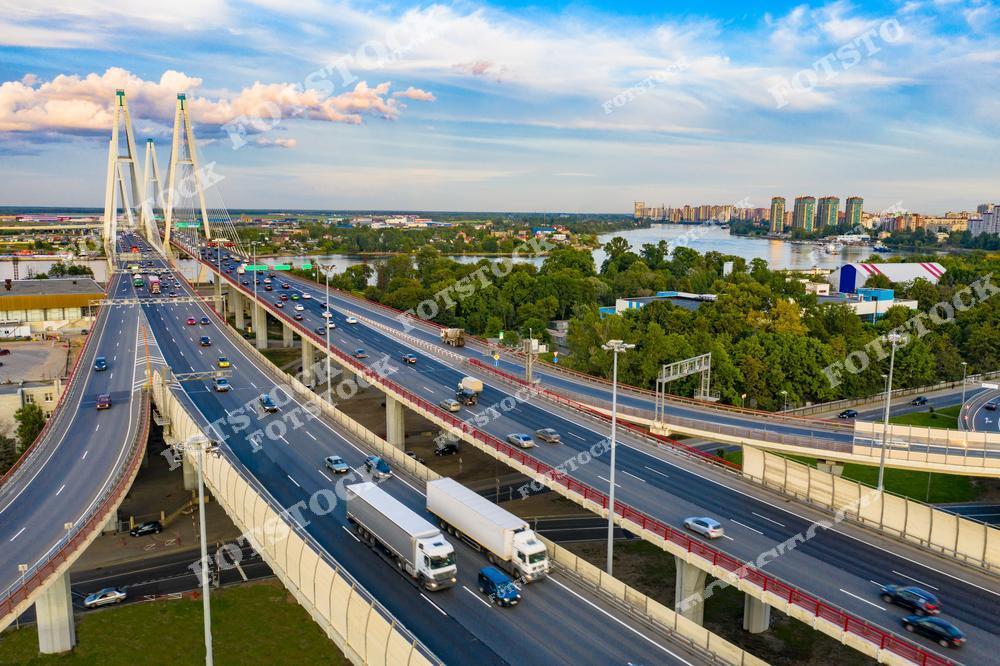 На фотографии изображен Вантовый мост, пересекающий реку Неву в Санкт-Петербурге. Движение автомобилей по многополосной магистрали демонстрирует транспортную активность города. На заднем плане видны жилые дома и зеленые массивы, создающие гармонию между урбанистикой и природой. Этот снимок подчеркивает развитую инфраструктуру и современный облик северной столицы.